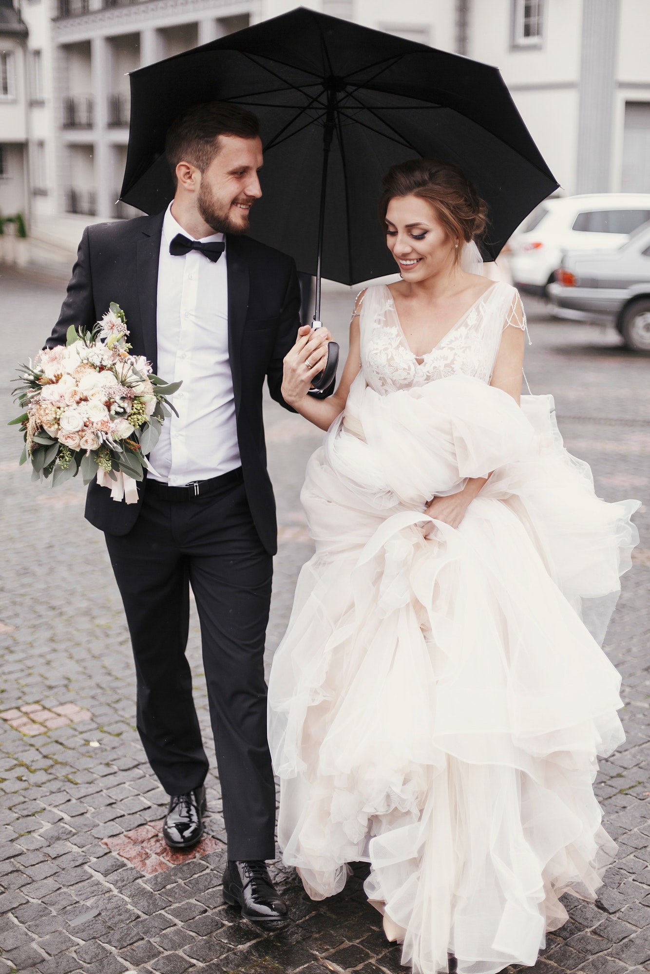 gorgeous-bride-and-stylish-groom-walking-under-umbrella-in-rainy-street-and-smiling.jpg